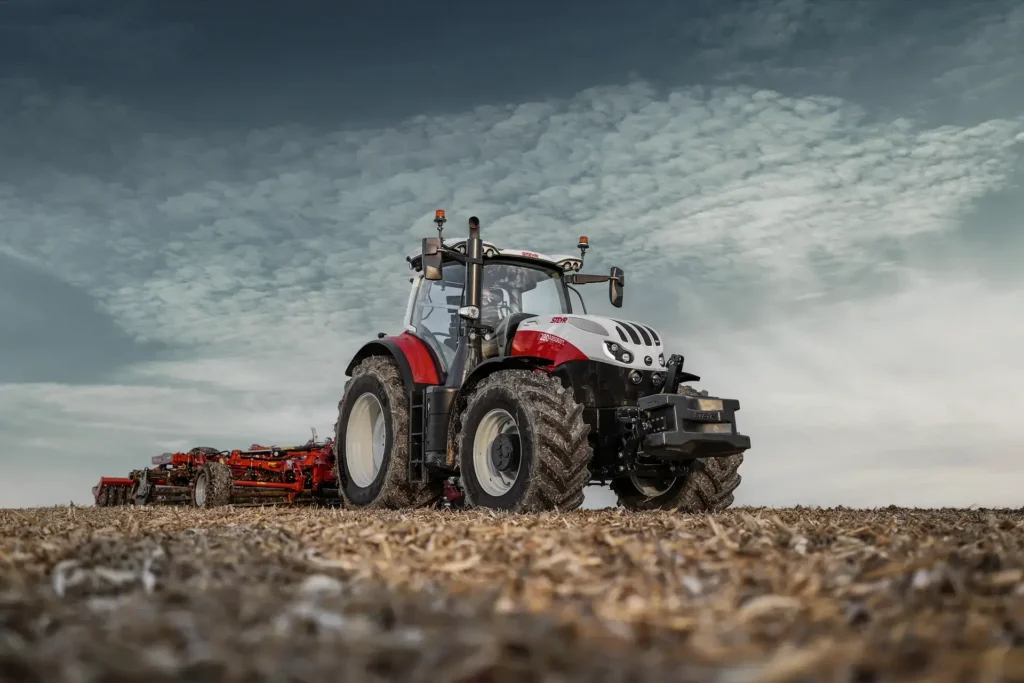 En Steyr 6280 Absolut CVT traktor trekker et rødt redskap på en pløyd åker, med desaturerte farger.