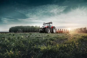 En Steyr Absolut CVT-traktor jobber på en åker i solnedgang. Traktoren er utstyrt med en plog og omringet av frodig grønt gress, med en tett skog i bakgrunnen. Sollyset som bryter gjennom horisonten skaper en dramatisk og stemningsfull scene.