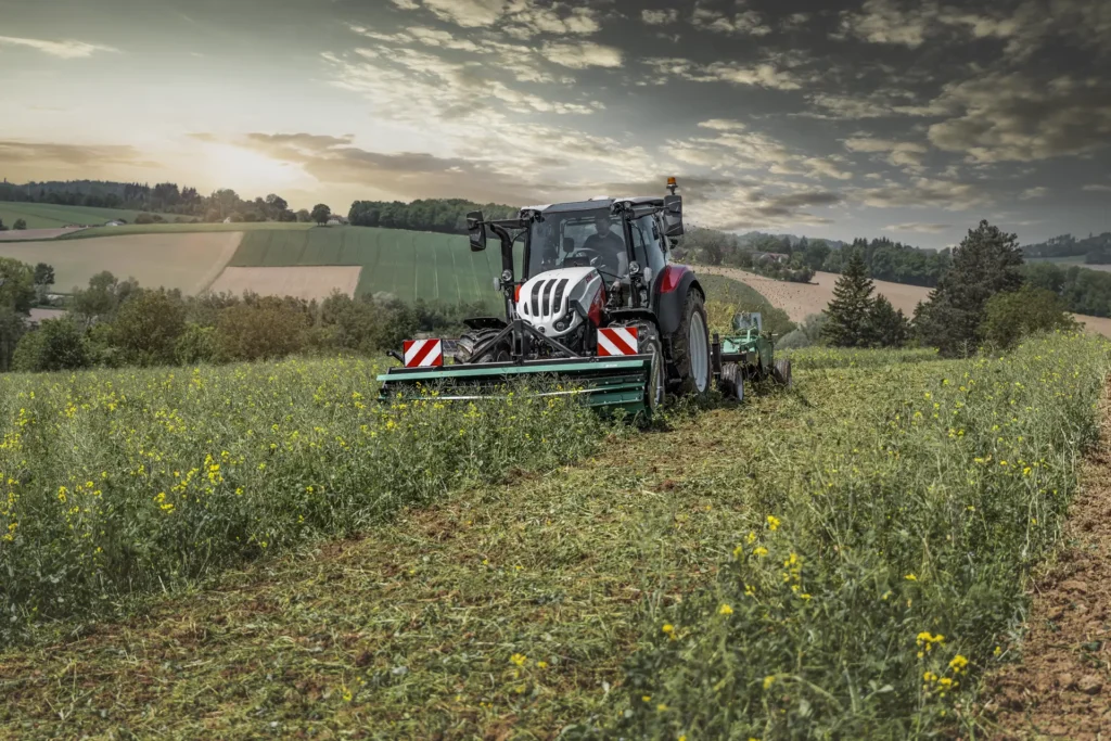 En Steyr-traktor klipper gress med en frontmontert slåmaskin i et grønt felt.