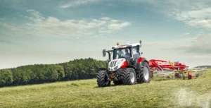 En Steyr Expert-traktor slår gress på en grønn eng med trær og blå himmel i bakgrunnen, og viser sin effektivitet for høsting og vedlikehold.