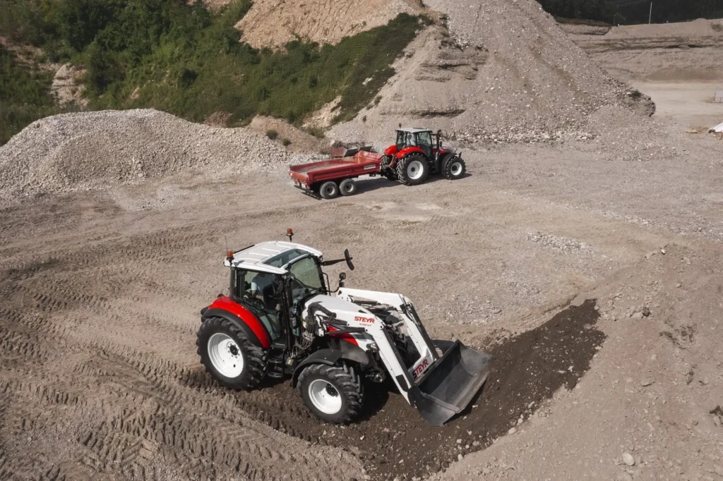 En STEYR traktor med frontlaster laster grus, mens en annen traktor med tilhenger står i bakgrunnen.