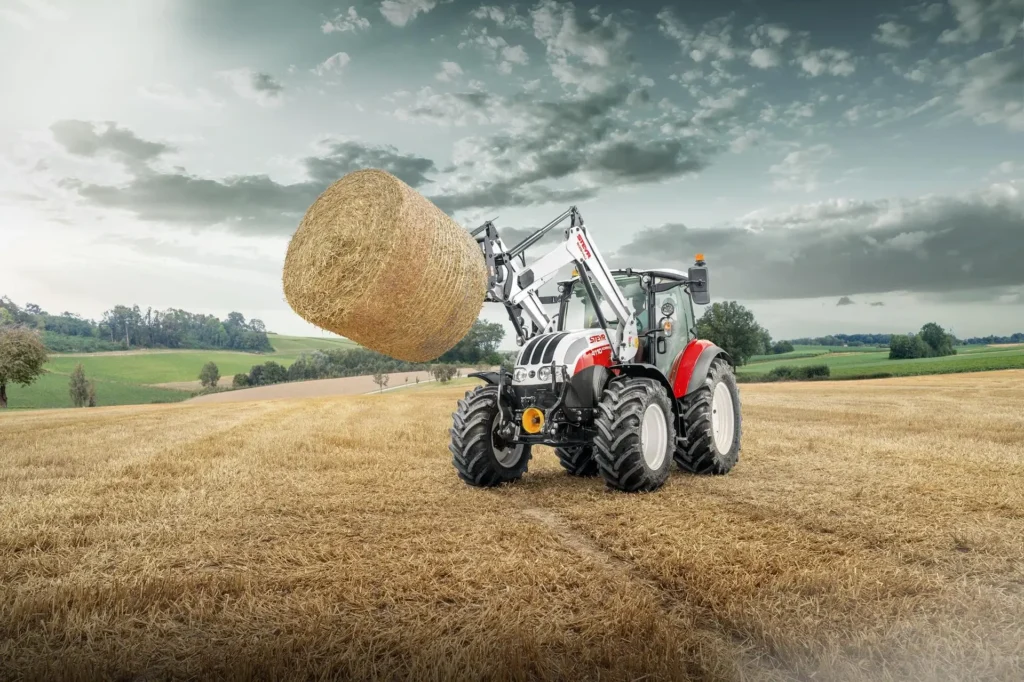 En STEYR traktor løfter en stor rundballe med frontlaster på et jorde, omgitt av vakker natur.