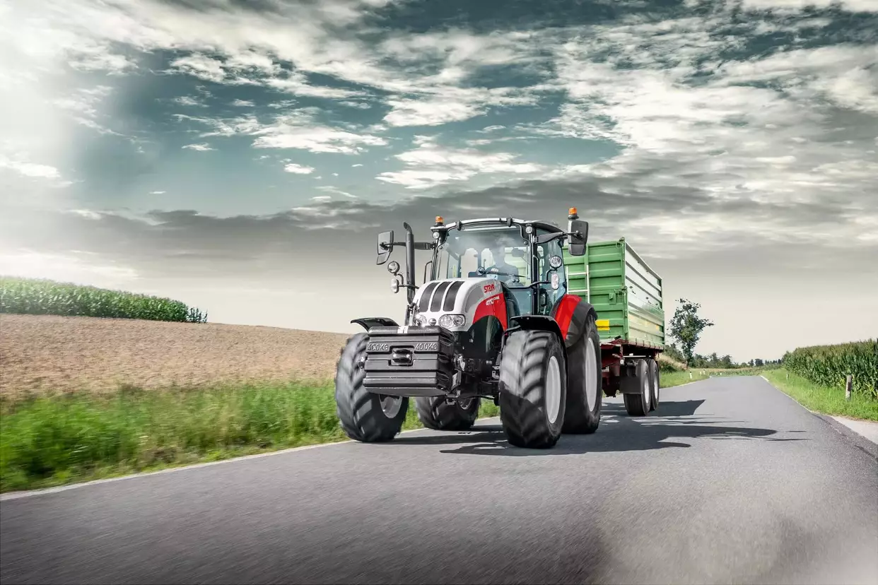 En Steyr-traktor med tilhenger på en landevei, omgitt av et landlig landskap.