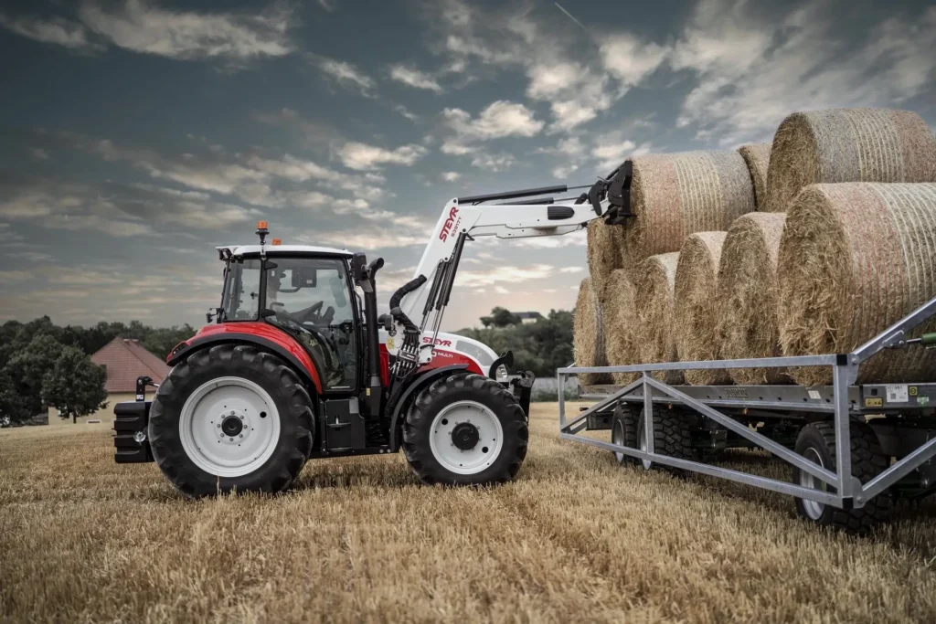 STEYR traktor med frontlaster løfter en rundballe opp på en ballehenger.