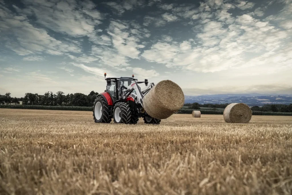 STEYR traktor med frontlaster transporterer en rundballe på en åker i solnedgang.