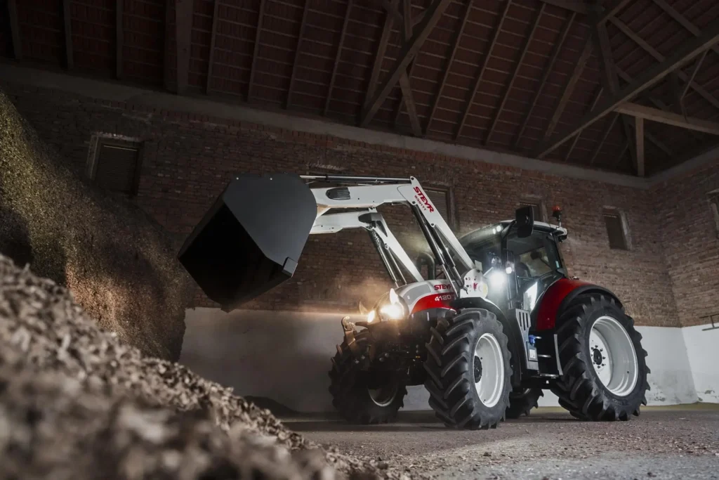 STEYR traktor med frontlaster jobber innendørs i et fjøs, med frontskuffen løftet.