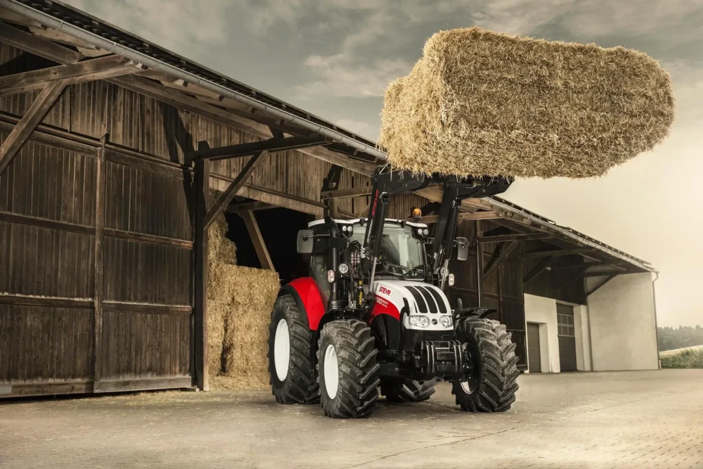 Steyr 4120 Multi-traktor med frontlaster som løfter en høyball foran en låve.