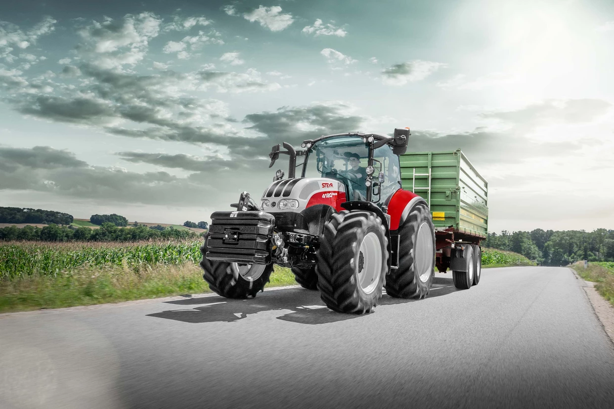 Steyr 4120 Multi traktor kjører på en landevei med tilhenger i et landlig landskap.