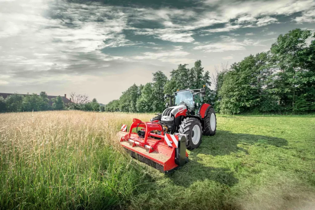 Steyr Multi traktor med beitepusser arbeider på et jorde omgitt av trær.