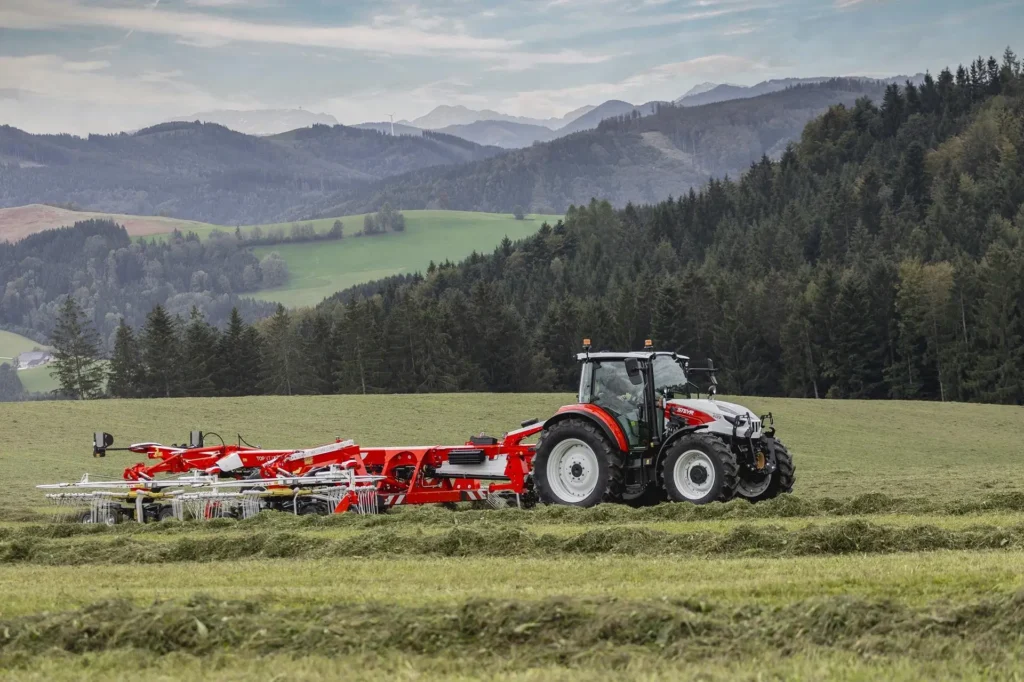 Steyr Plus-traktor i drift med en stor høyvender, omgitt av fjellandskap.