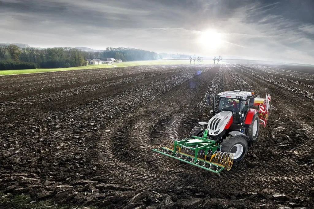 Steyr Profi traktor arbeider på en oppdyrket åker med såmaskin, soloppgang i bakgrunnen.