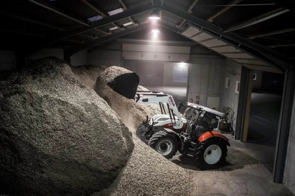 Steyr Profi traktor laster store mengder flis eller sagflis inne i en lagerhall med frontlaster.