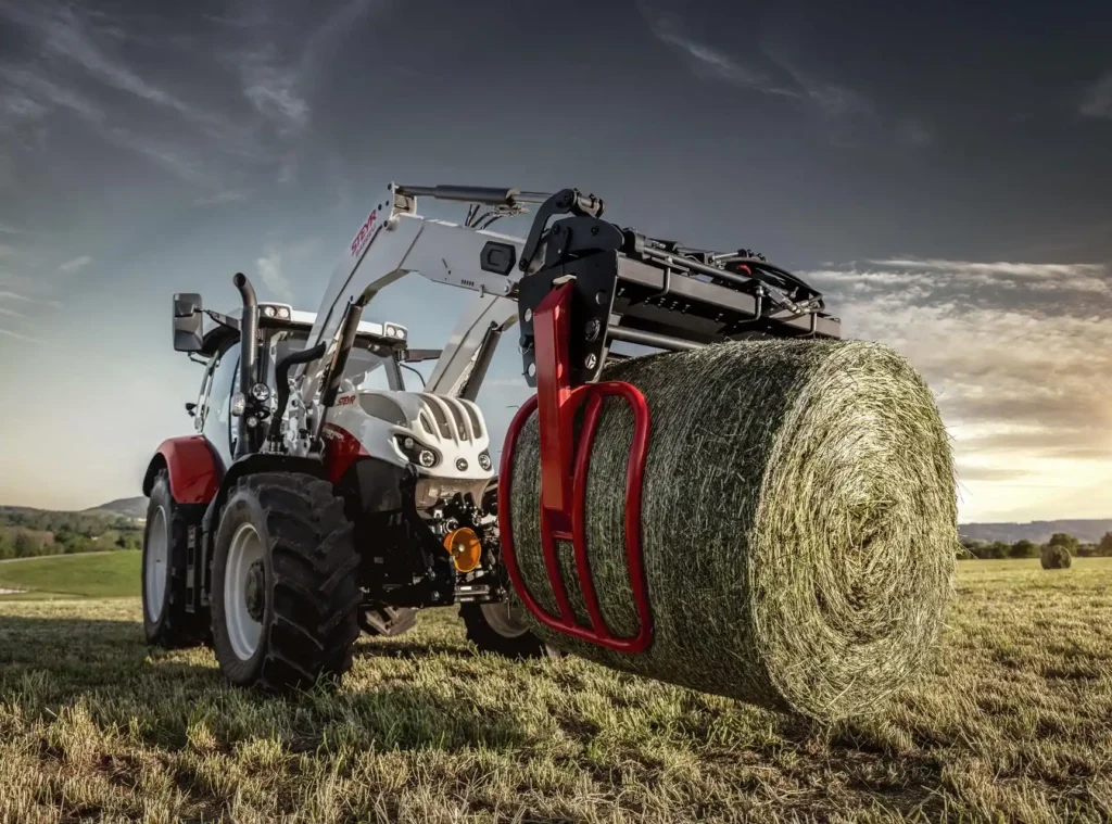 Steyr Profi traktor utstyrt med en rundballeklemme løfter en stor høyball på en åker i skumringen.