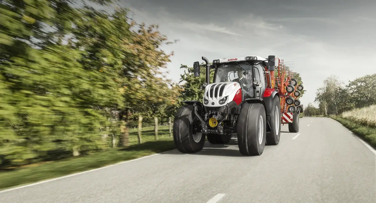 En Steyr Profi 6145 traktor i transportmodus på en landevei, med en stor landbruksmaskin festet bak.