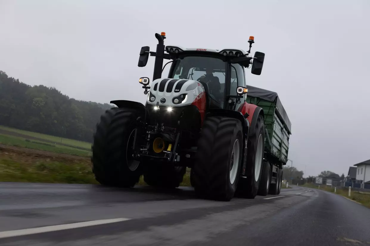 Steyr 6280 Absolut CVT kjører på en landevei med en grønn tilhenger, i overskyet vær.