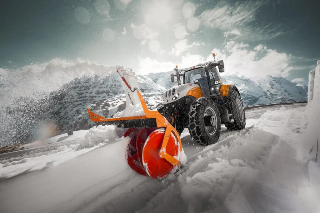 Steyr Terrus traktor med snøfreser fjerner snø i et vinterlandskap med fjell i bakgrunnen.