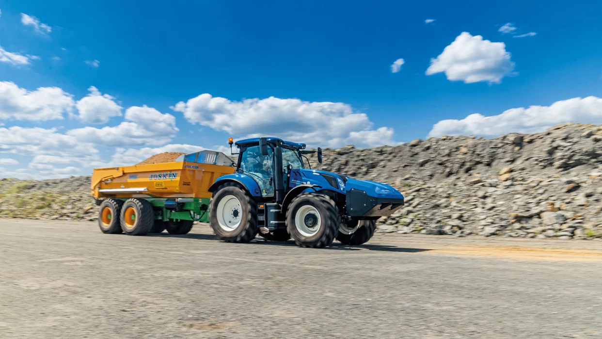 En blå New Holland traktor trekker en tung lastehenger over en steinete vei, med fokus på robusthet og ytelse.