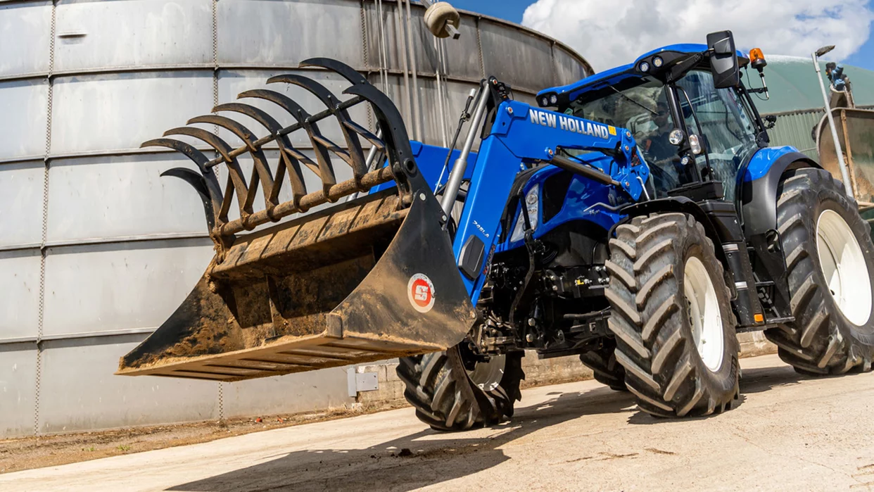 En New Holland traktor med en stor gripelaster foran et industrielt anlegg, klar for tungt arbeid.