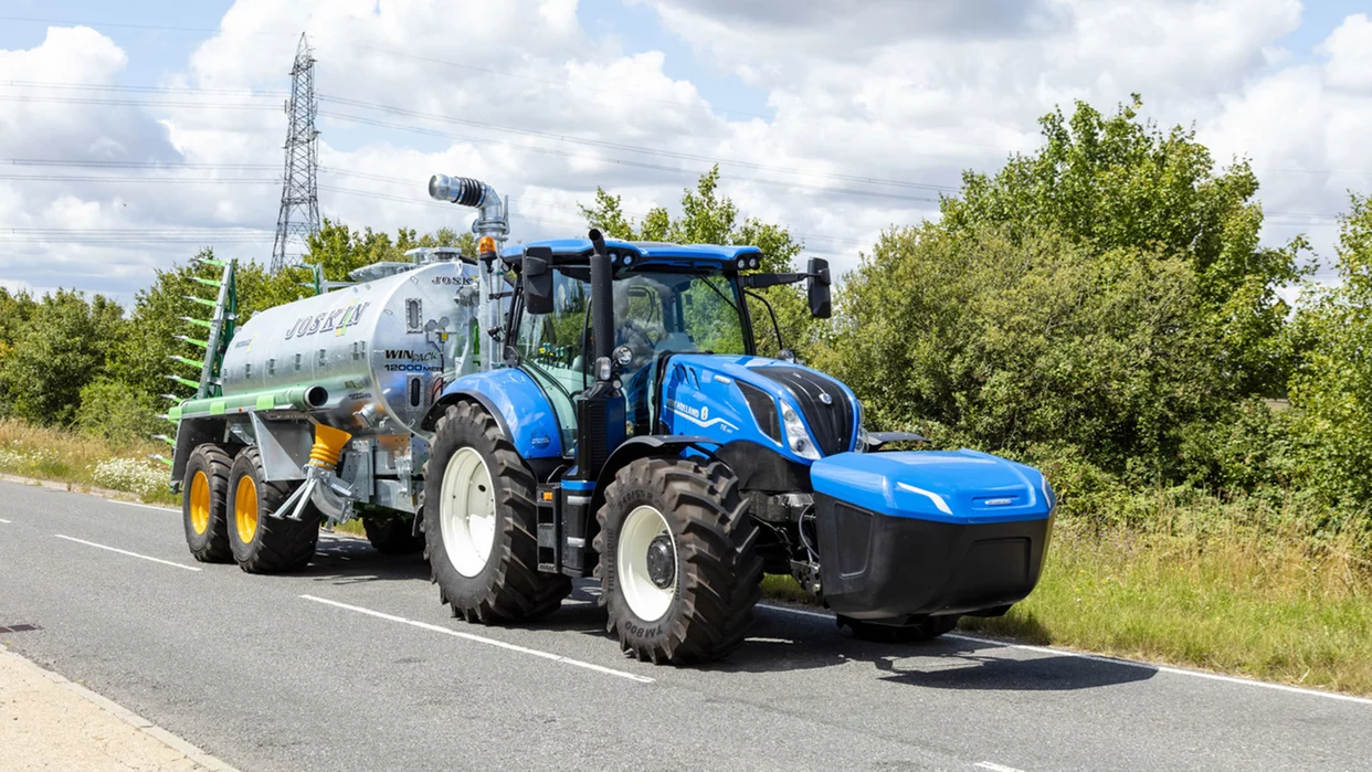 En blå New Holland traktor med tilhenger som tømmer last i et åpent område, under en klar blå himmel.