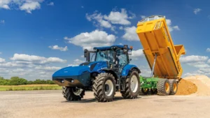 En blå New Holland traktor med tilhenger som tømmer last i et åpent område, under en klar blå himmel.