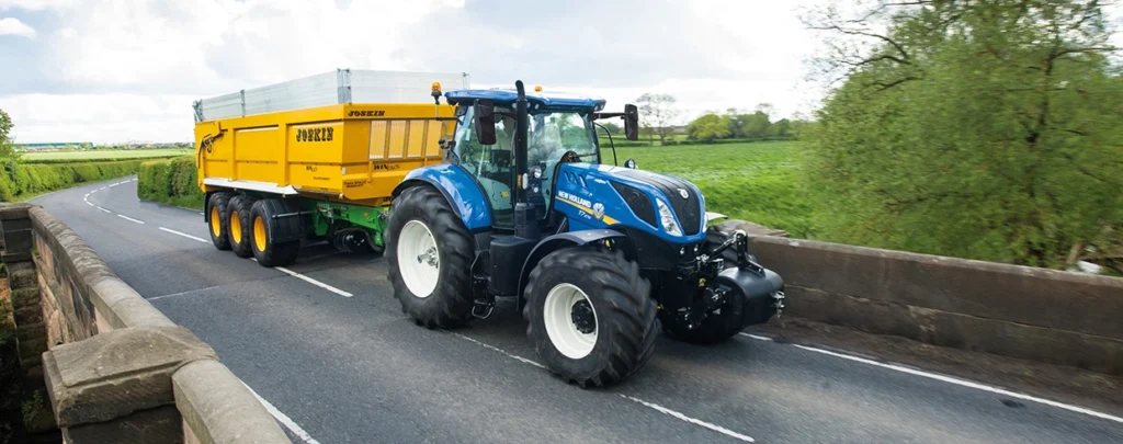 En blå New Holland T7 LWB-traktor som trekker en gul JOSKIN-henger over en bro, som demonstrerer robusthet og allsidighet.