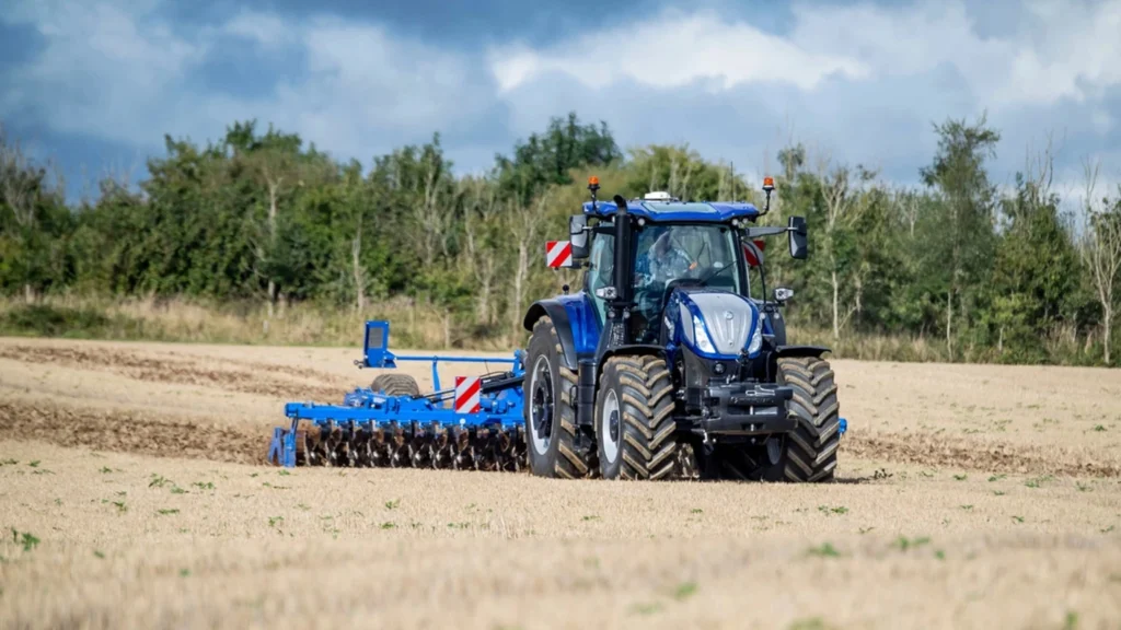 En New Holland T7 LWB-traktor som trekker en gul presse over et felt i dagslys, som fremhever effektivitet i landbruksarbeid.