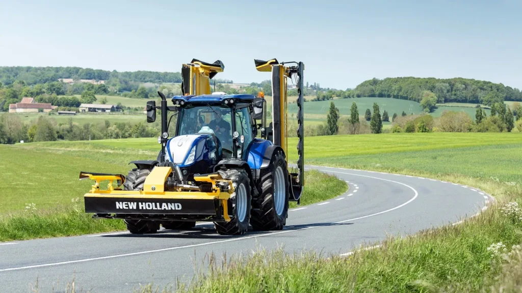 New Holland-traktor på en landevei omgitt av grønne åkre og åser.