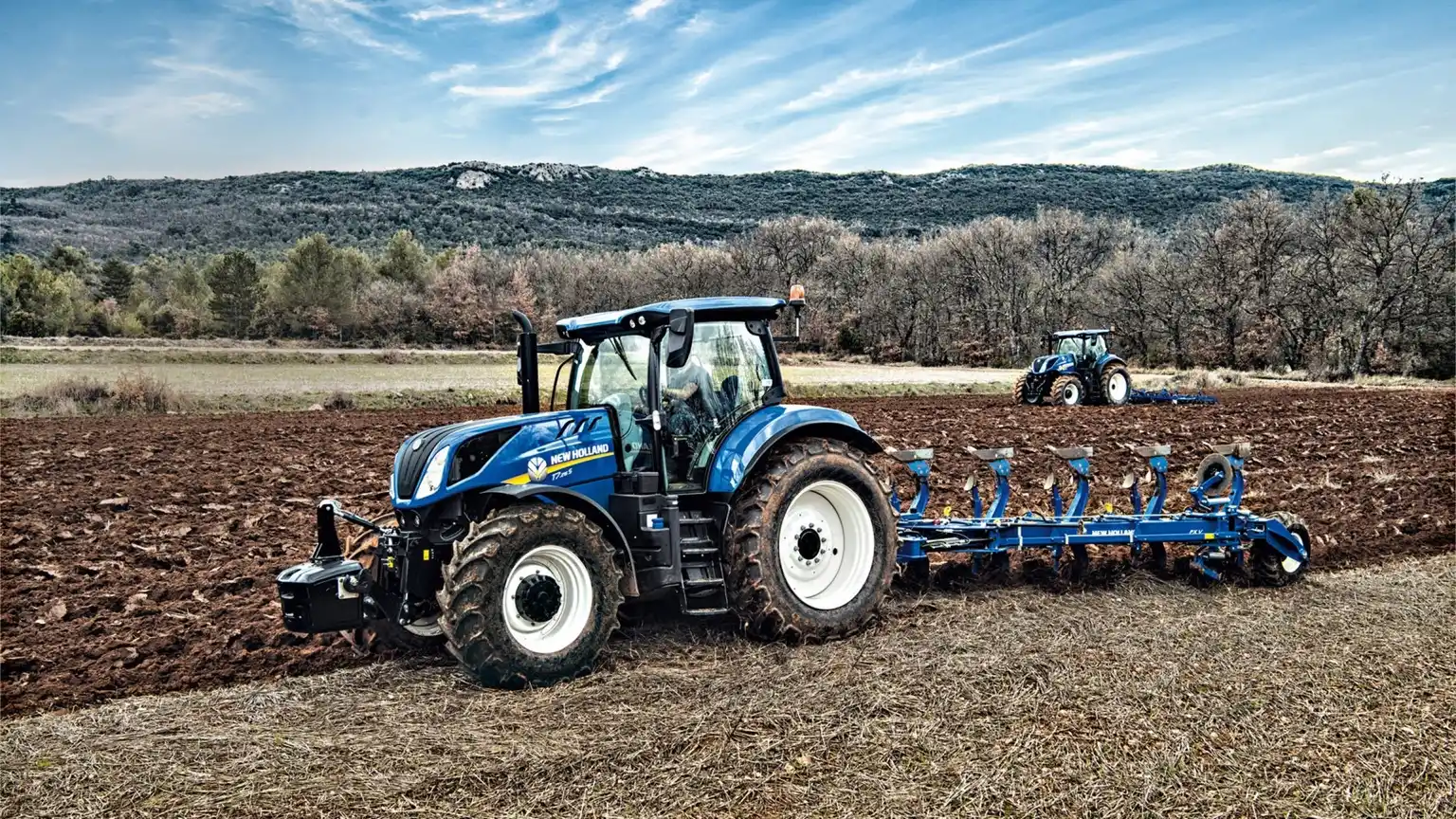 En New Holland T7 traktor som pløyer en åker, med et naturskjønt fjell- og skoglandskap i bakgrunnen under en klar blå himmel.